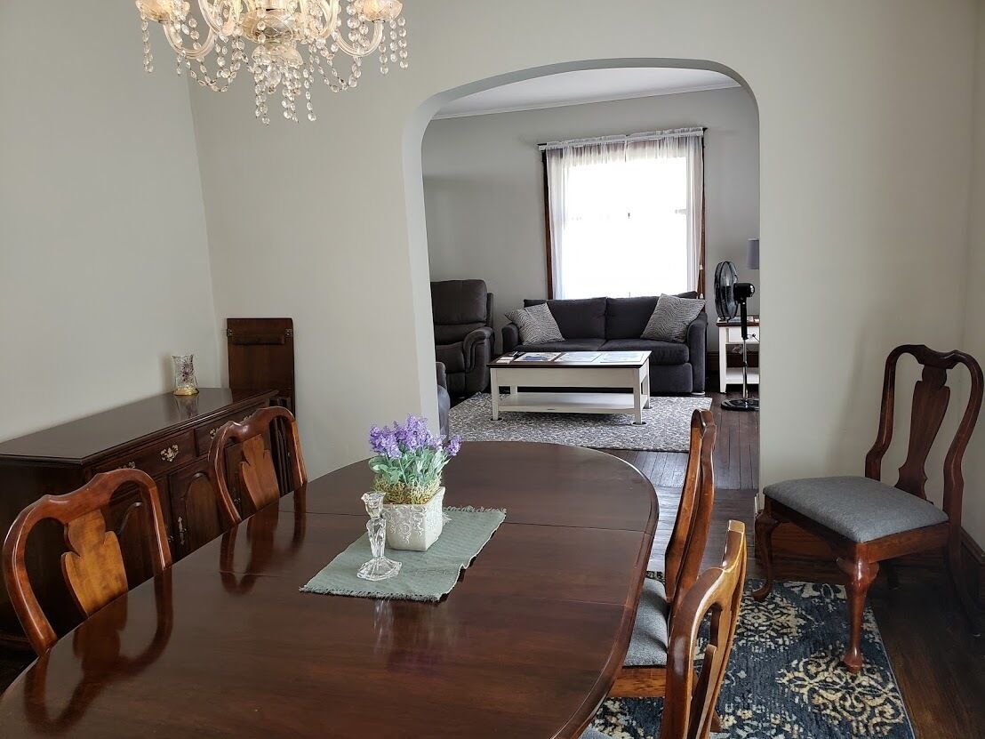 A dining area with a wooden table, chairs, and a chandelier. There is a sofa and a window in the background.