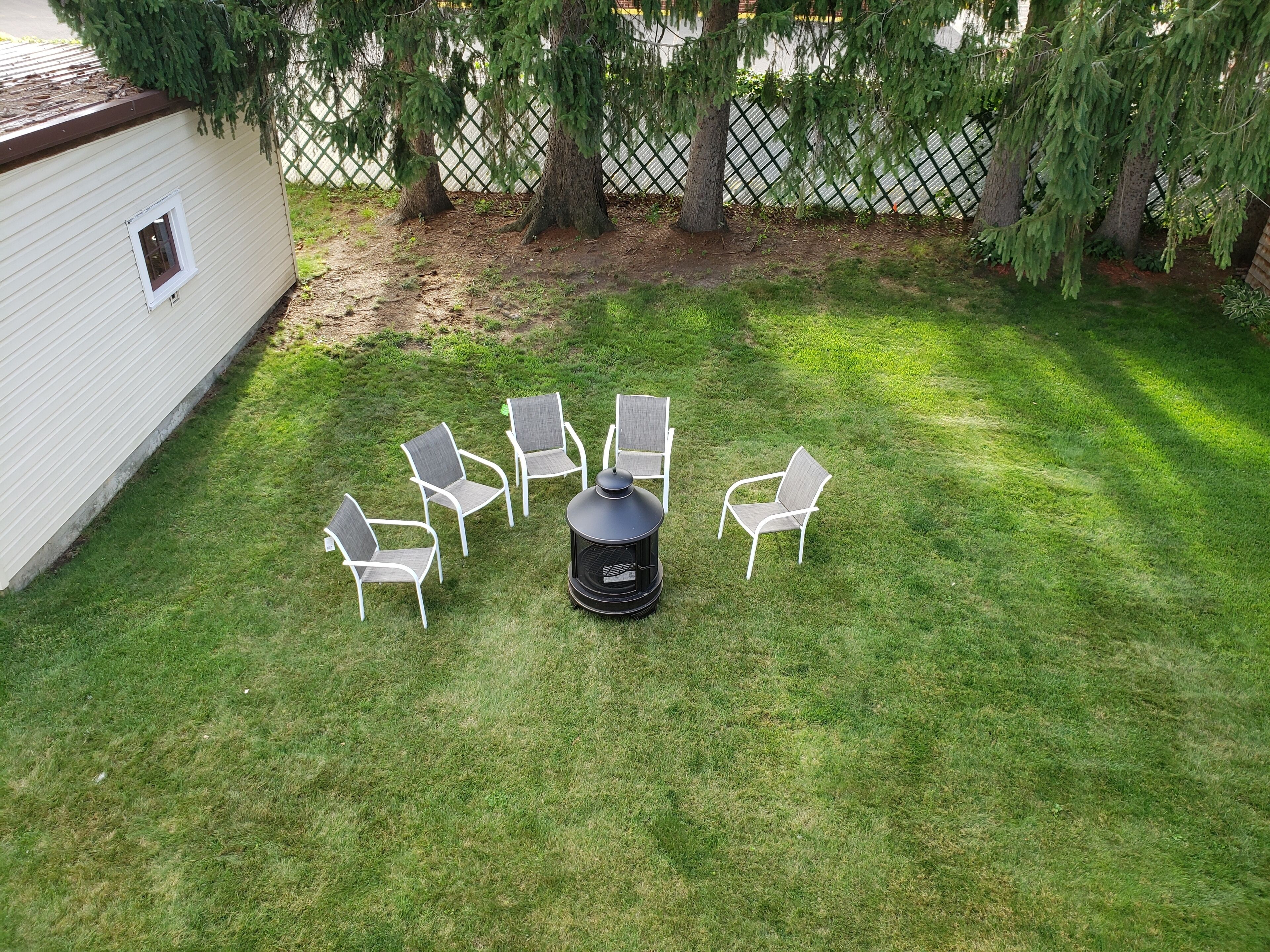 Ein Garten mit einem weißen Gebäude, einem schwarzen Grill und fünf weißen Stühlen, die darum angeordnet sind.
