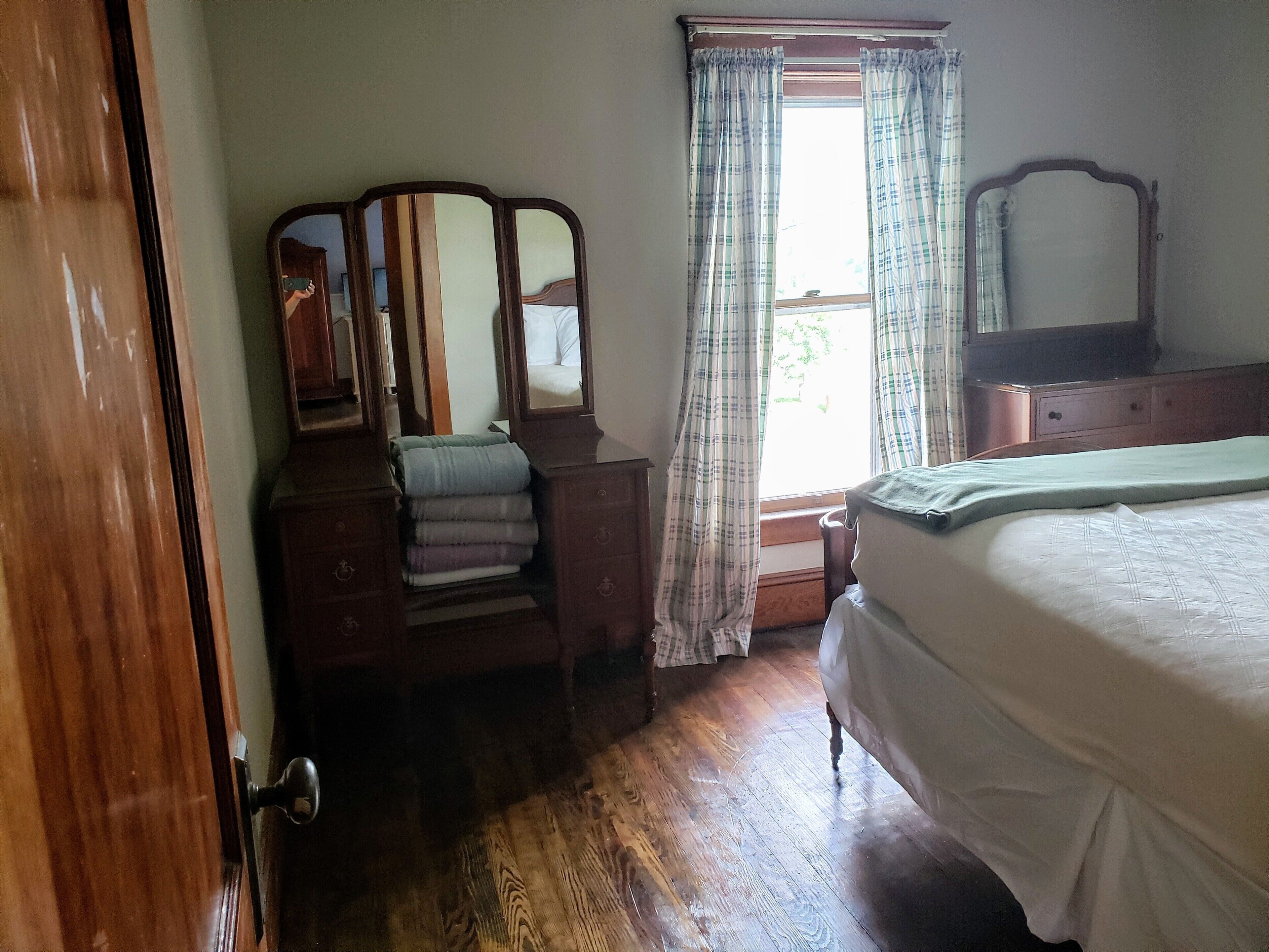 Une chambre à coucher avec une commode en bois, un lit avec une literie blanche, un miroir et une fenêtre avec des rideaux.