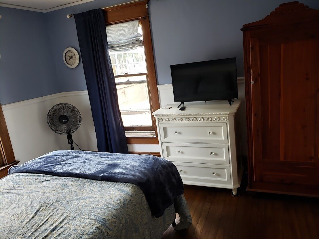 A bedroom with a bed, a dresser, a television, a fan, and a window with curtains.