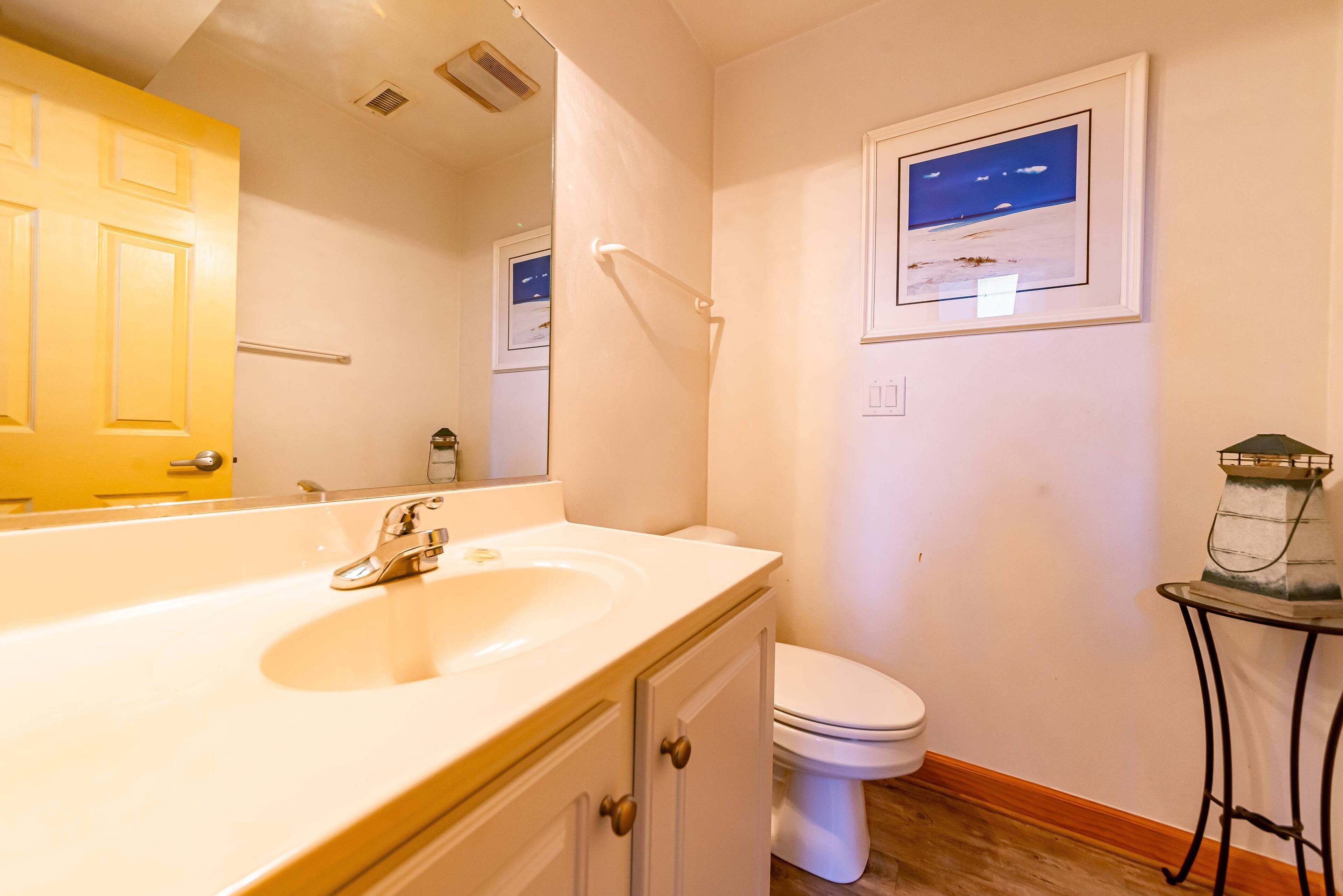 Une salle de bain avec un lavabo blanc, des toilettes, un tableau encadré au mur et une petite table sur laquelle est posé un objet décoratif.