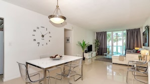 A modern living room with a dining area, a TV, and a large window with curtains. - Beachfront Condo | Ocean Breeze & Resort Pool (Hollywood)