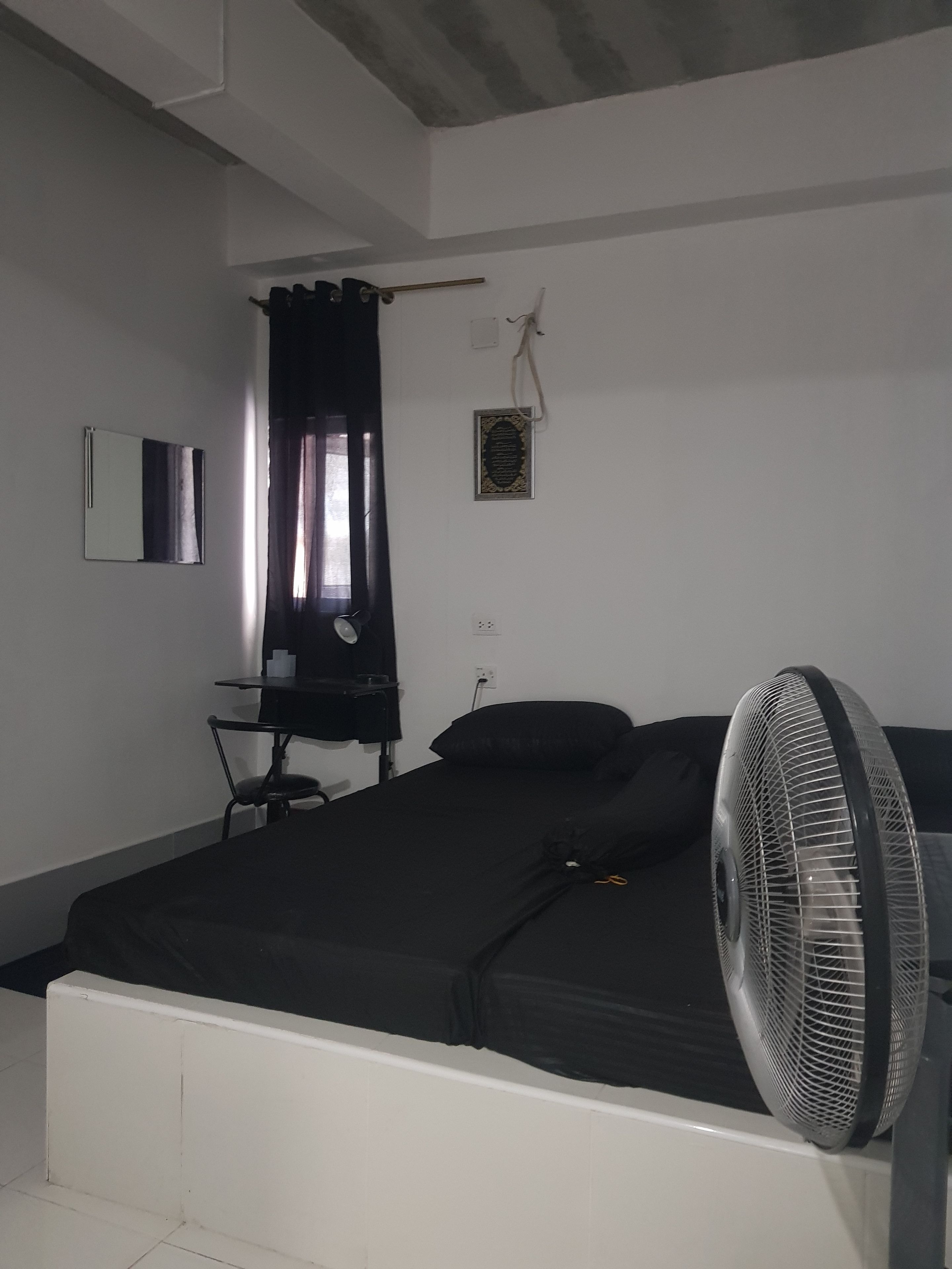 Quarto pequeno e simples com cama, escrivaninha, cadeira, ventilador e janela com cortinas.
