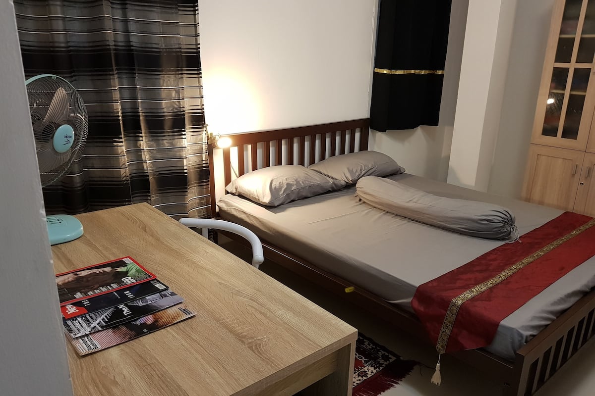 A bedroom with a wooden bed, a bedside table with magazines, a standing fan, and a nightstand.