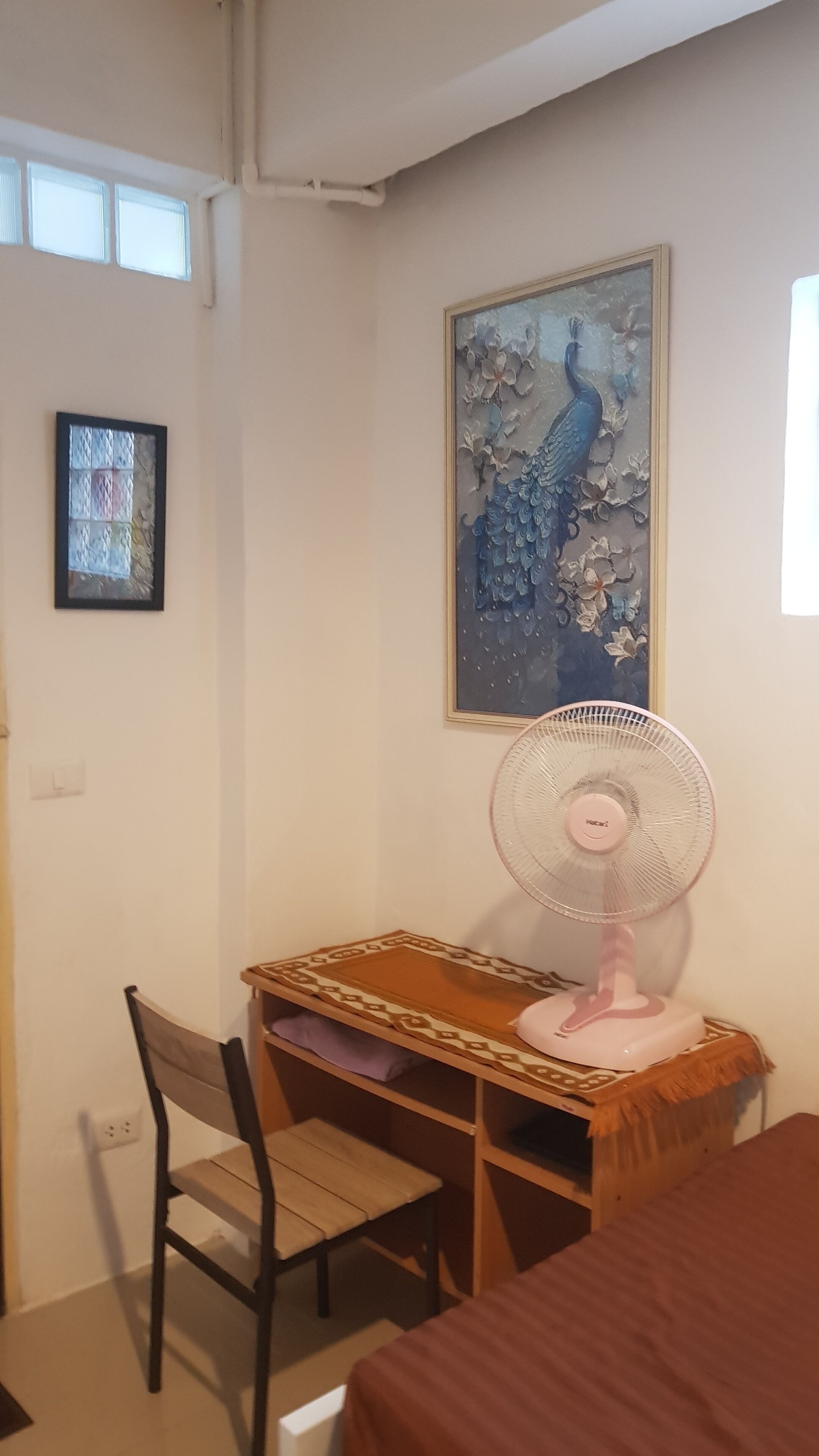 A room with a desk, chair, bed, and a wall-mounted fan.