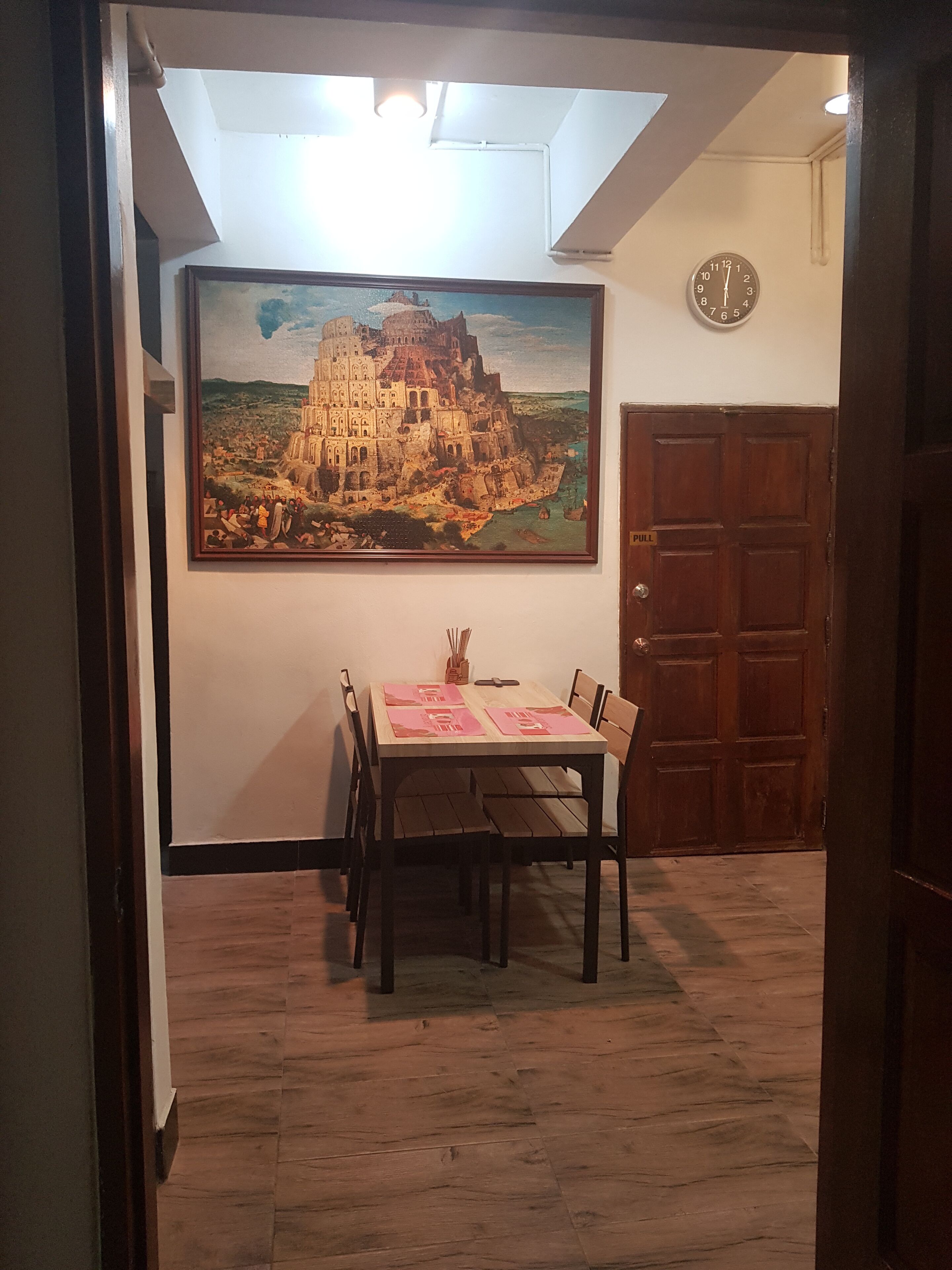 A dining area with a table set for two, a painting of a historical building on the wall, and a clock.