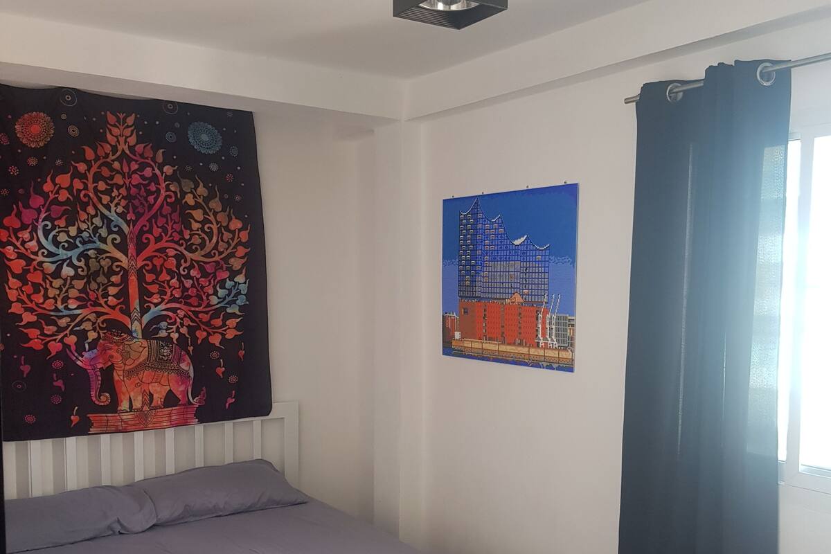 A bedroom with a bed, a wall tapestry, a framed picture, a window with curtains, and a white trash bin.