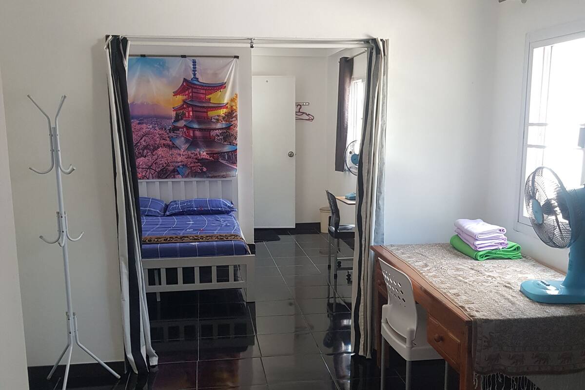 A hotel room with a bed, a desk with a chair, a standing fan, and a window.