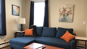 Ein Wohnzimmer mit einem blauen Sofa, zwei orangefarbenen Kissen, einem Holztisch, einer Lampe, einem Blumenbild und einem Fenster mit dunklen Vorhängen.