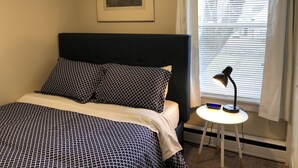 Ein Schlafzimmer mit einem Bett, einem Nachttisch, einer Lampe, einem Fenster mit Jalousien und einem gerahmten Bild an der Wand.