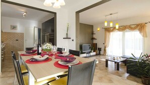 Un espace de repas moderne avec une grande table dressée pour quatre personnes, un téléviseur en arrière-plan et un coin salon avec un canapé et des plantes.