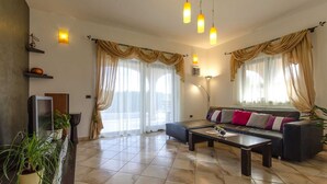 Un salon avec un canapé, une table basse et un téléviseur posé sur un meuble. On y trouve des rideaux, une plante et un luminaire fixé au mur.