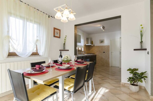 A dining area with a table set for four, chairs, a chandelier, and a view into the kitchen.