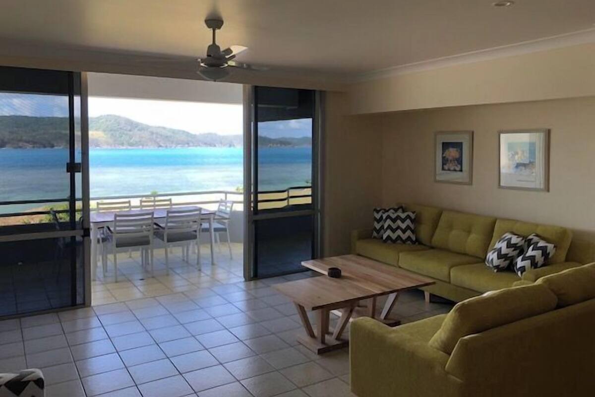 A living room with a sofa, a coffee table, and a dining area with chairs and a table. There is a large window offering a view of the sea and hills.