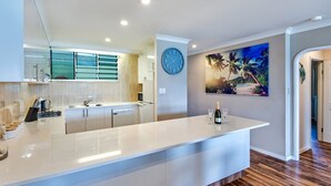 A modern kitchen with a central island, a clock on the wall, and a beach-themed painting. - The Elite F208 by HamoRent on Hamilton Island (Hamilton Island)