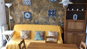 Una sala acogedora con pared de piedra, un sofá amarillo, cojines estampados, una mesa de comedor de madera y un aparador con una planta pequeña.