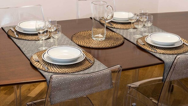 Uma mesa de jantar posta com copos de vidro transparente, pratos brancos e um jarro de vidro transparente, tudo envolto por jogos americanos estampados.