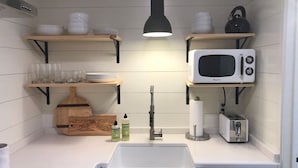 A compact kitchen with a white sink, a microwave, a toaster, and wooden shelves above the counter.
