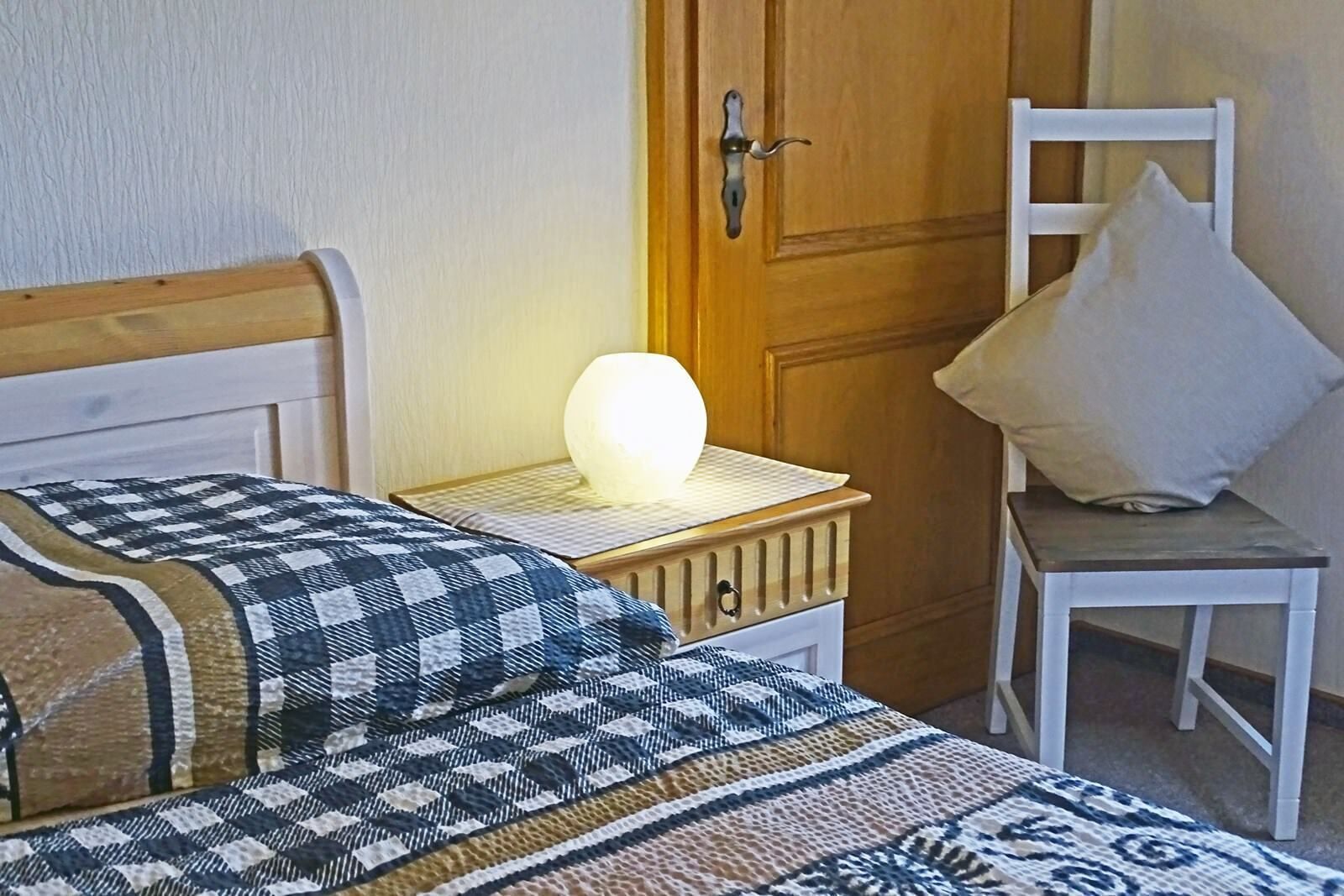 Quarto com duas camas, mesa de cabeceira com abajur, uma cadeira e porta de madeira.