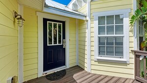 A yellow house with a black front door, a window with shutters, and a porch with a doormat.