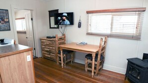 Une pièce chaleureuse avec une table à manger en bois, des chaises, un téléviseur fixé au mur, un petit poêle noir et une fenêtre avec des stores.
