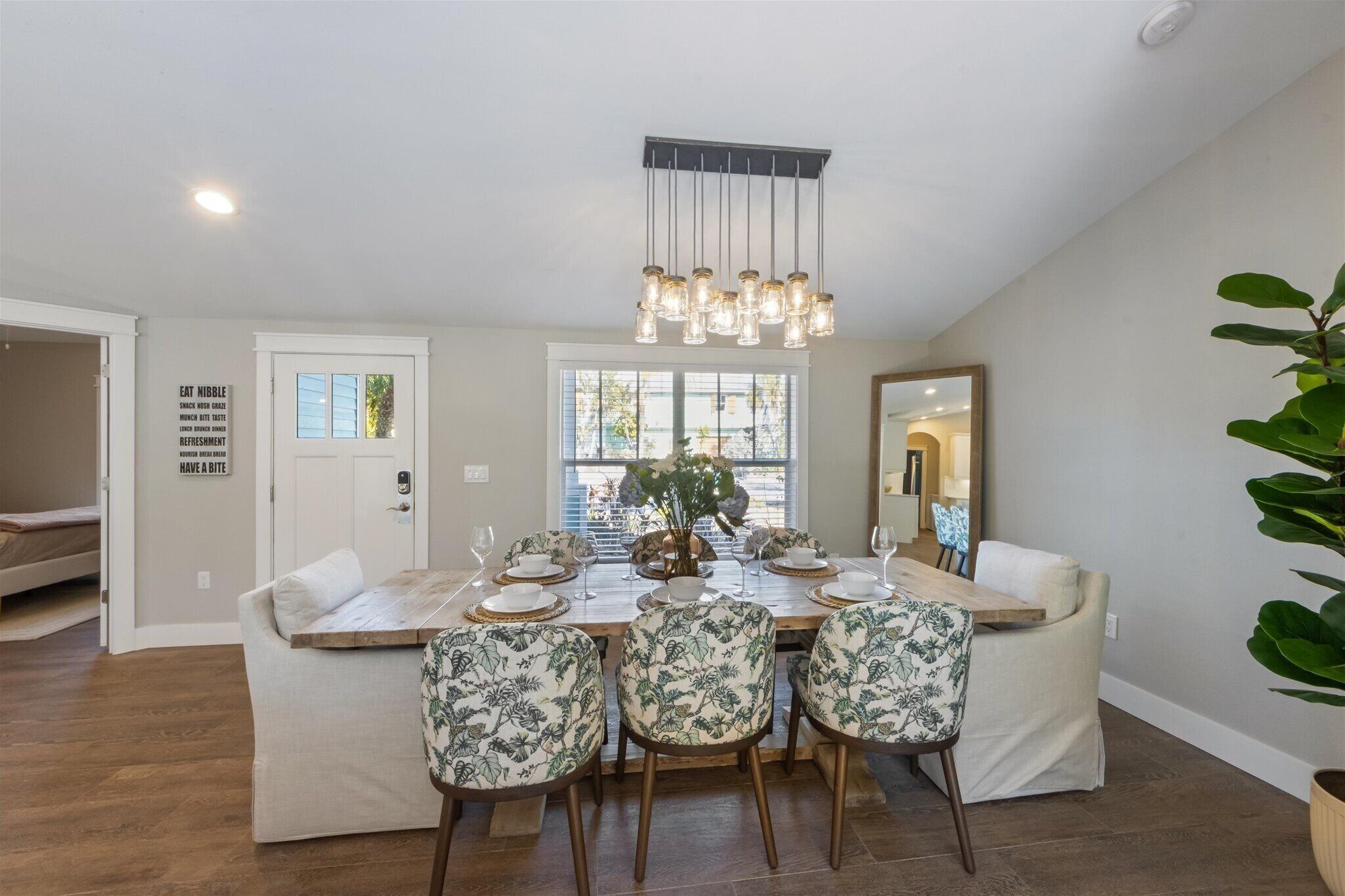 A dining area with a wooden table set for four, surrounded by floral-patterned chairs. A hanging light fixture is above the table, and a large mirror is on the wall to the right.