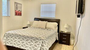 A bedroom with a bed, two bedside tables, a ceiling fan, a window with blinds, and a wall-mounted TV.