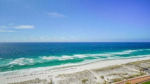 A beach with clear blue water, a sandy shore, and a road with parked cars.