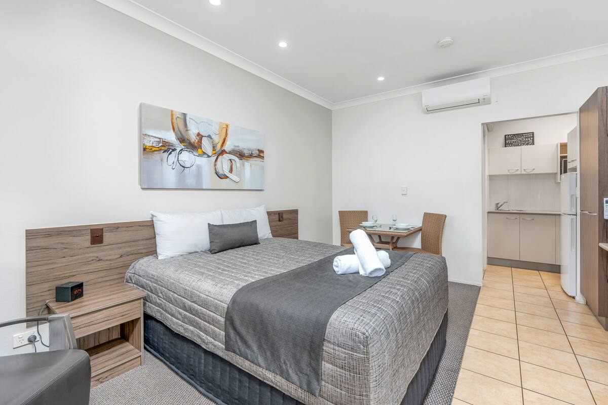 Ein modernes Hotelzimmer mit einem großen Bett, einem Nachttisch, einem Schreibtisch und einem Sessel. An der Wand hängt ein Bild und über dem Schreibtisch befindet sich eine Klimaanlage.