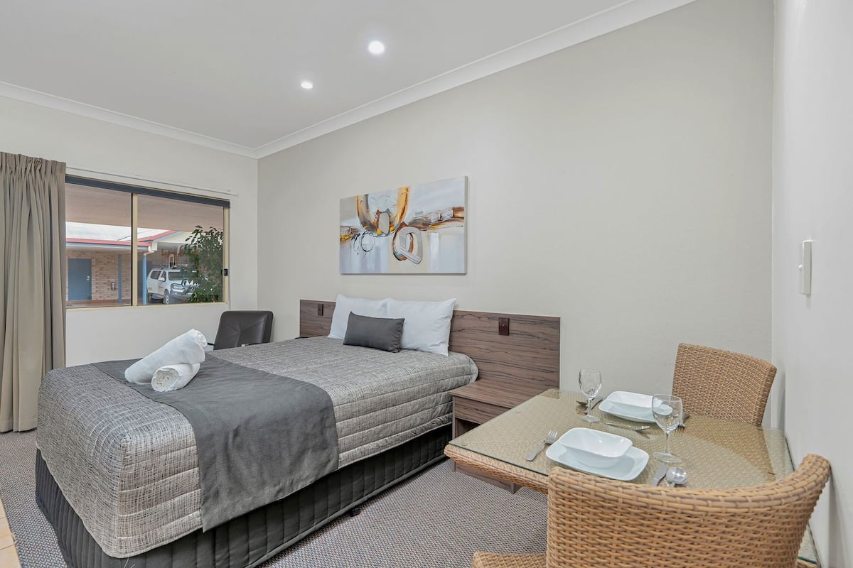 Quarto de hotel com cama, mesa de jantar e cadeiras, um quadro na parede e janela com cortinas.