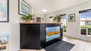 A reception desk with a digital screen displaying 'Midlander' in orange letters, framed artwork on the walls, and potted plants. - Midlander Motor Inn (Emerald)