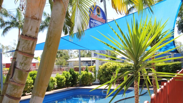 A swimming pool surrounded by palm trees and a blue shade sail. - Midlander Motor Inn (Emerald)