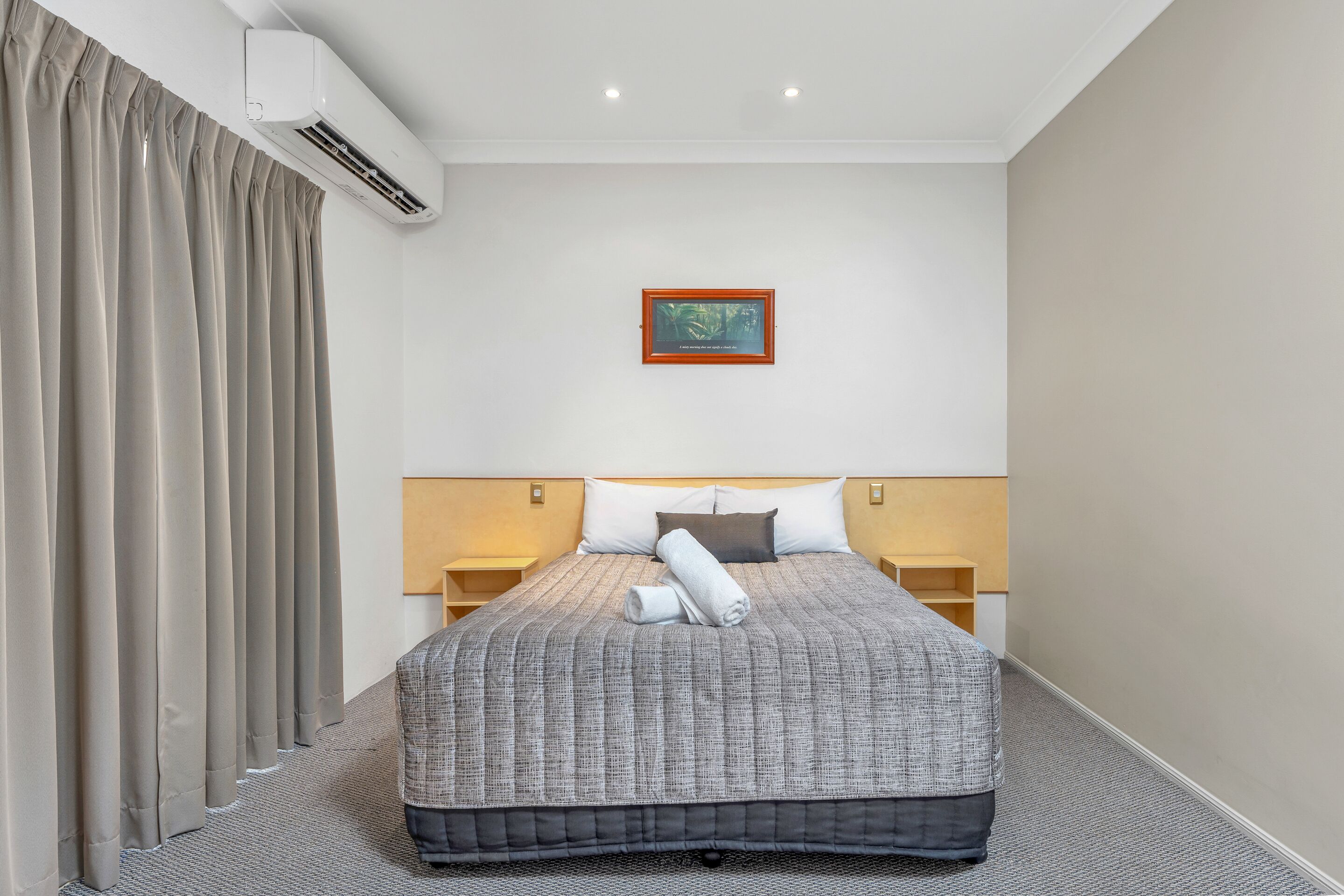 A hotel room with a large bed, a headboard with a built-in shelf, a wall-mounted air conditioner, and a framed picture on the wall.