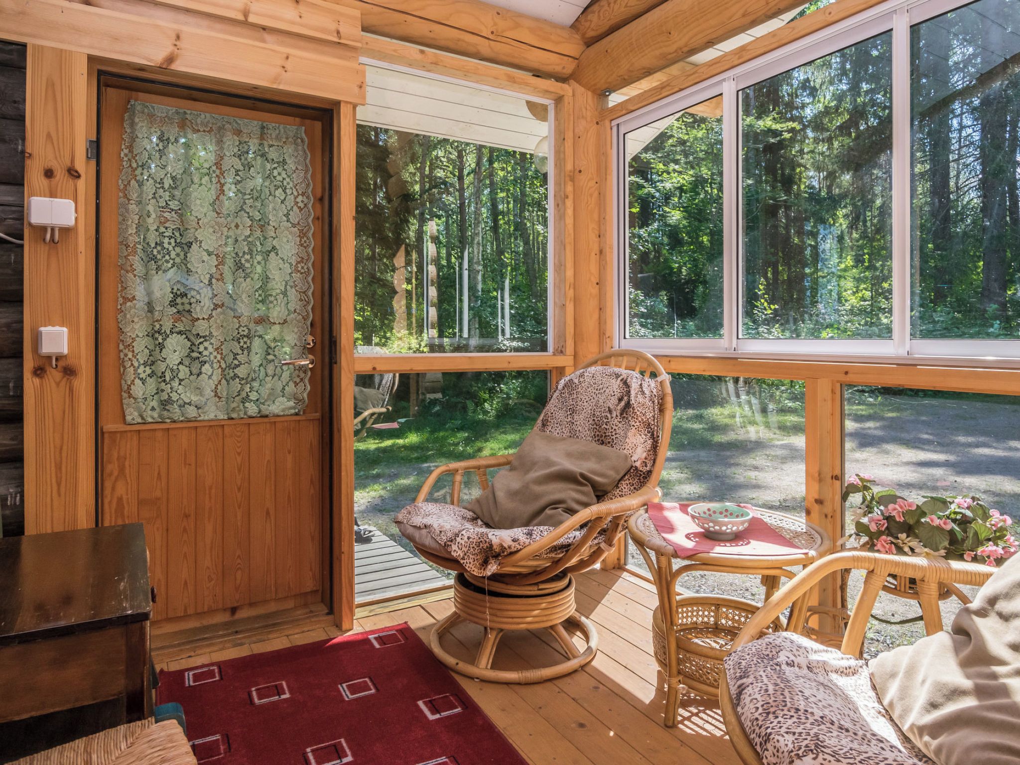 Interior de una cabaña de madera con una mecedora, una mesa de comedor y vistas a un bosque a través de amplias ventanas.