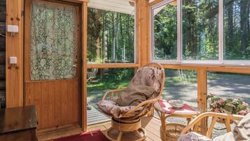 Interior de una cabaña de madera con una mecedora, una mesa de comedor y vistas a un bosque a través de amplias ventanas.