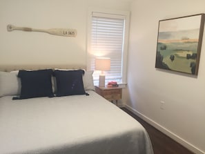 Un dormitorio con cama, almohadas, una mesita de noche con lámpara, una ventana con persianas y un cuadro en la pared.