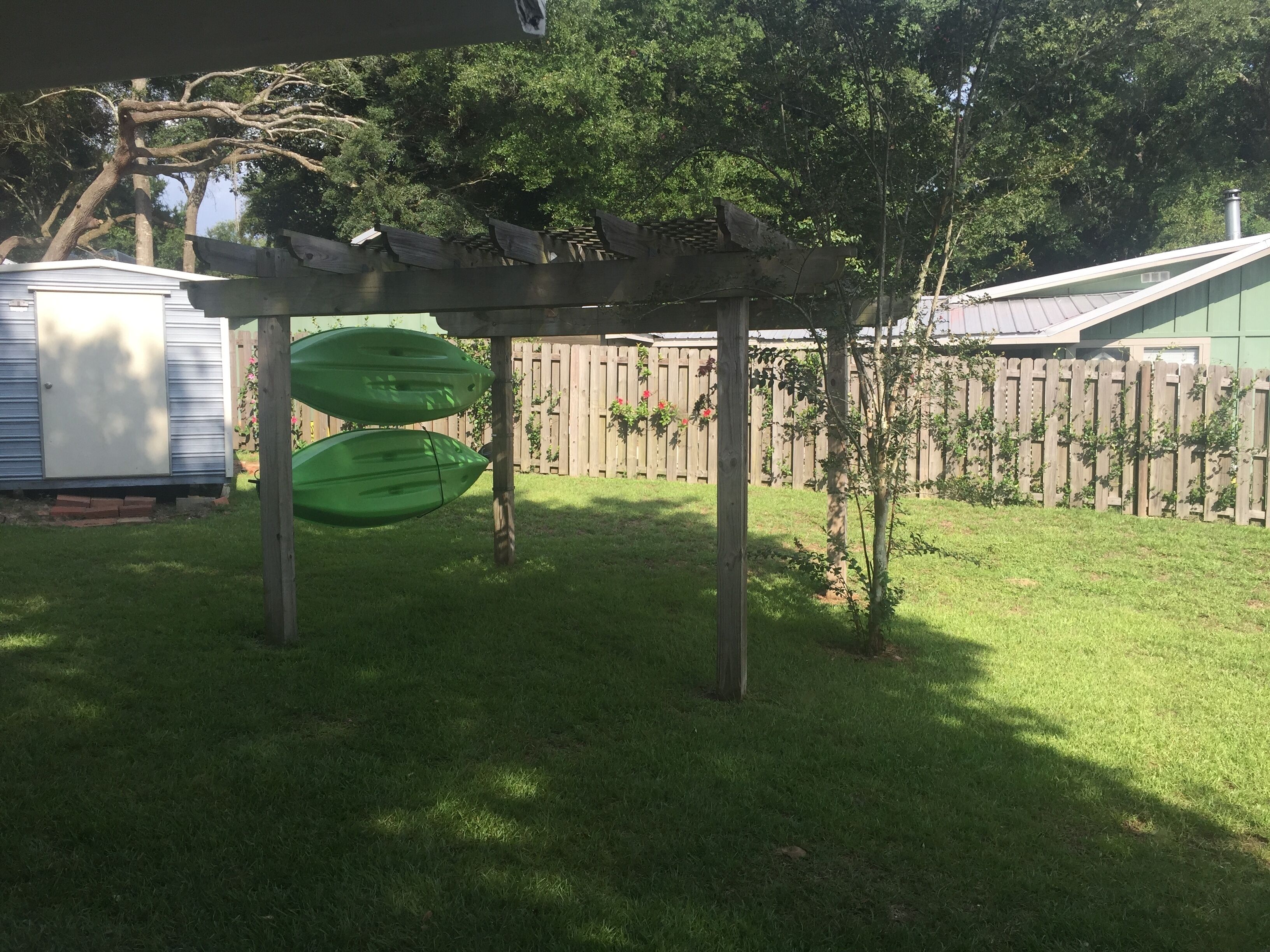 Un patio trasero con un pérgola de madera, dos kayaks verdes, una casa rodante blanca y una valla de madera.