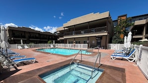 A swimming pool with lounge chairs and umbrellas, surrounded by a fenced area with a building in the background. - LUXURY CONDO GROUND FLOOR, STEPS TO POOL, 2 BEDROOMS, 1 BATH, SLEEPS 5 (Osoyoos)