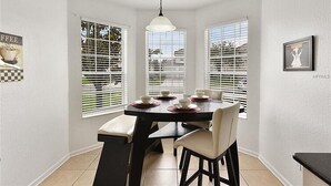 Comedor con mesa dispuesta para cuatro personas, paredes blancas y ventanas grandes con persianas.