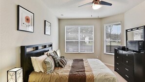 Un dormitorio con cama, ventilador de techo y dos ventanas con persianas.