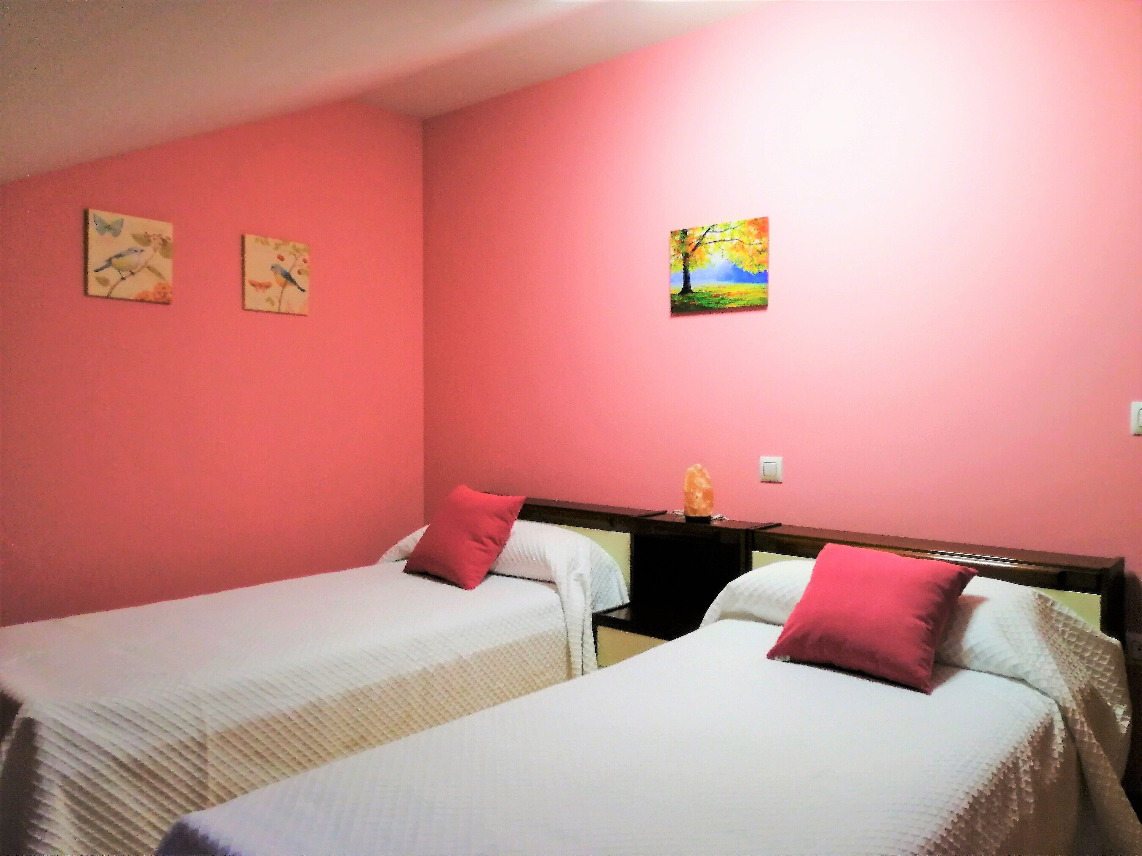 Une chambre avec deux lits, chacun étant équipé de draps blancs et d’un oreiller rouge. Deux tableaux ornent le mur, l’un représentant des oiseaux et l’autre, un paysage.