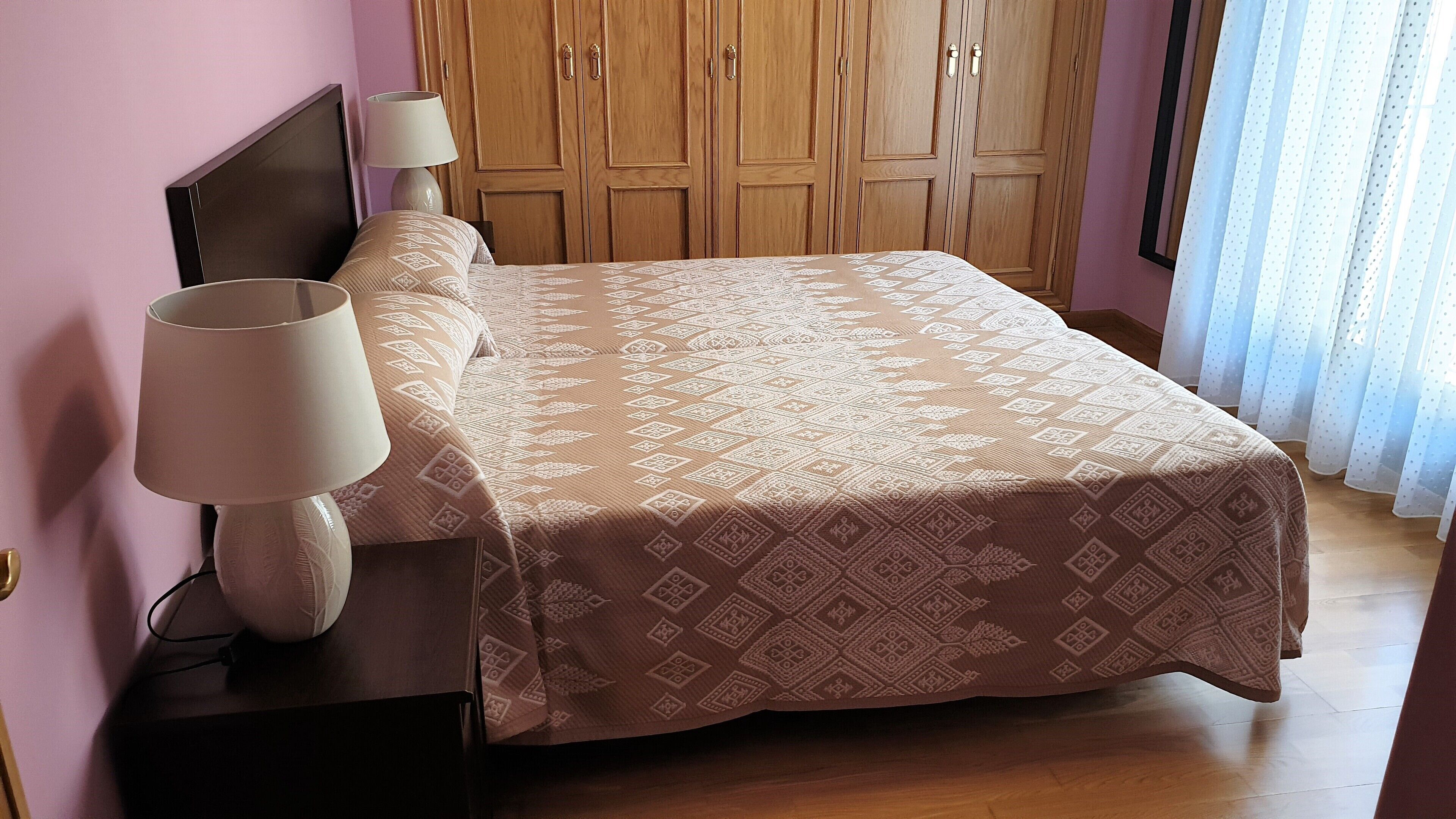 A neatly made bed with a patterned bedspread, a bedside lamp, and a wooden wardrobe in the background.
