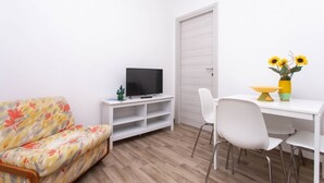Ein Zimmer mit einer Couch, einem Fernseher auf einem Schrank, einem Esstisch mit Stühlen und einer Tür.
