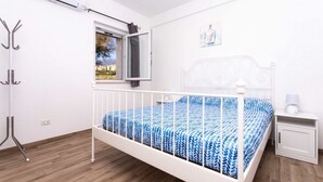 Ein Schlafzimmer mit einem weißen Bett, einem Fenster mit Vorhängen, einem Nachttisch mit Lampe und einem Kleiderständer.