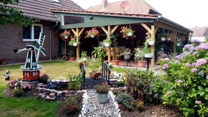 A house with a windmill, a covered porch with hanging flower pots, and a garden with various plants and flowers. - Lovingly furnished apartment is looking forward to young and old guests.  (Friedeburg)
