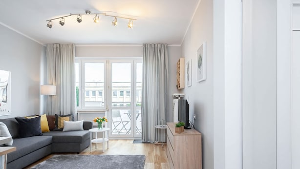 Una sala moderna con un sofá seccional gris, una pequeña mesa blanca con un jarrón de flores, una alfombra gris y un mueble de madera.