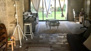 A stone-walled room with a stone floor, a sofa, a small table, a tripod, and a view of a garden through large windows. - Beautiful stone house in the village of Vers Pont du Gard (Vers-Pont-du-Gard)