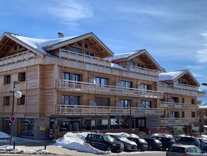 A multi-story wooden building with balconies, a ski slope visible in the background, and a parking area with cars. - 3-room apartment for 6 near slopes, Alpe d'Huez (Huez)
