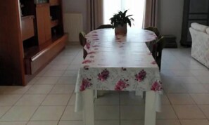 Une table à manger recouverte d’une nappe à motifs floraux, entourée de chaises, dans une pièce dont le sol est carrelé et qui est équipée d’un radiateur.