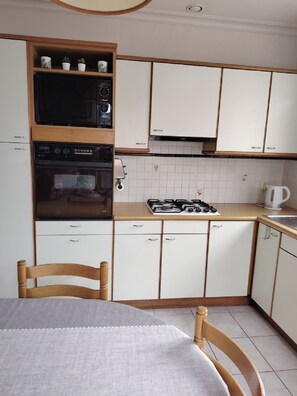 Une cuisine compacte avec des meubles blancs, un four à micro-ondes, un four traditionnel et une cuisinière à gaz. On y trouve une chaise en bois et une table recouverte d’une nappe blanche.